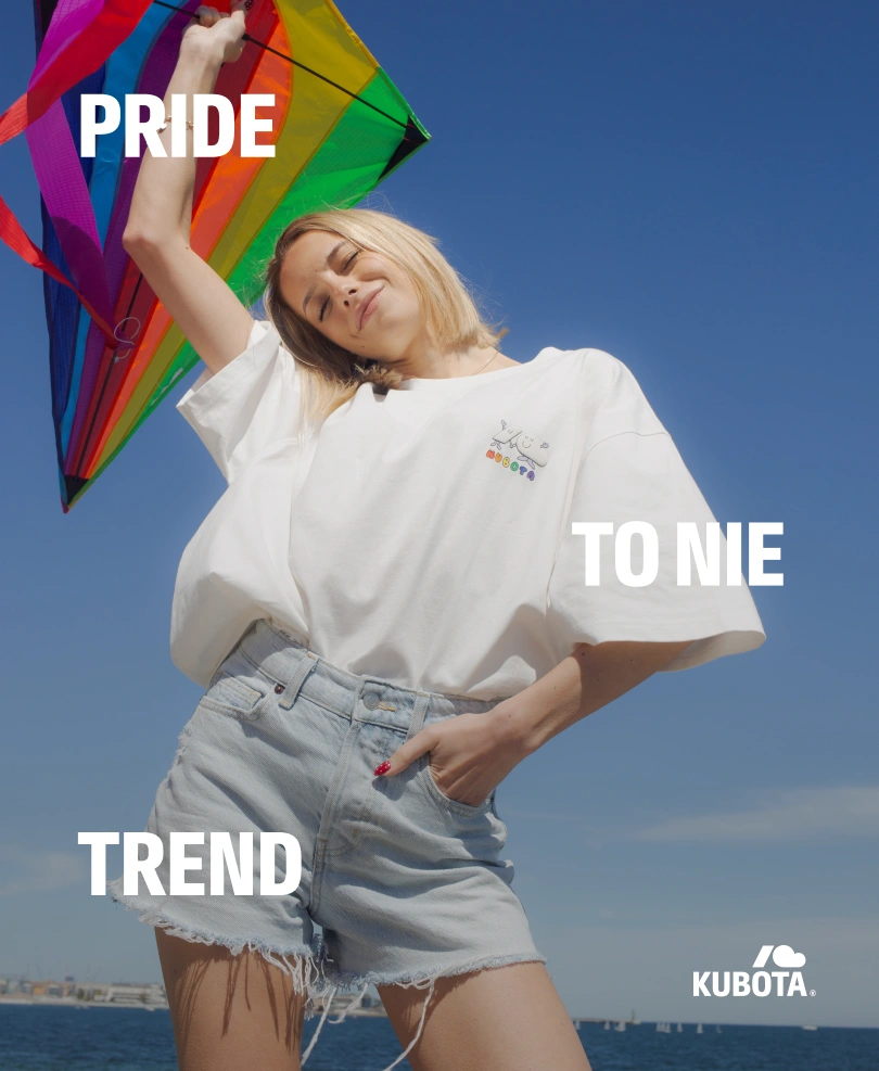 Grafika wizerunkowa prezentująca odzież z kategorii Pride Month. Kobieta w białym t-shircie Comfy Club Pride trzyma tęczowy latawiec na tle błękitnego nieba. Na grafice widnieje napis "Pride to nie trend".