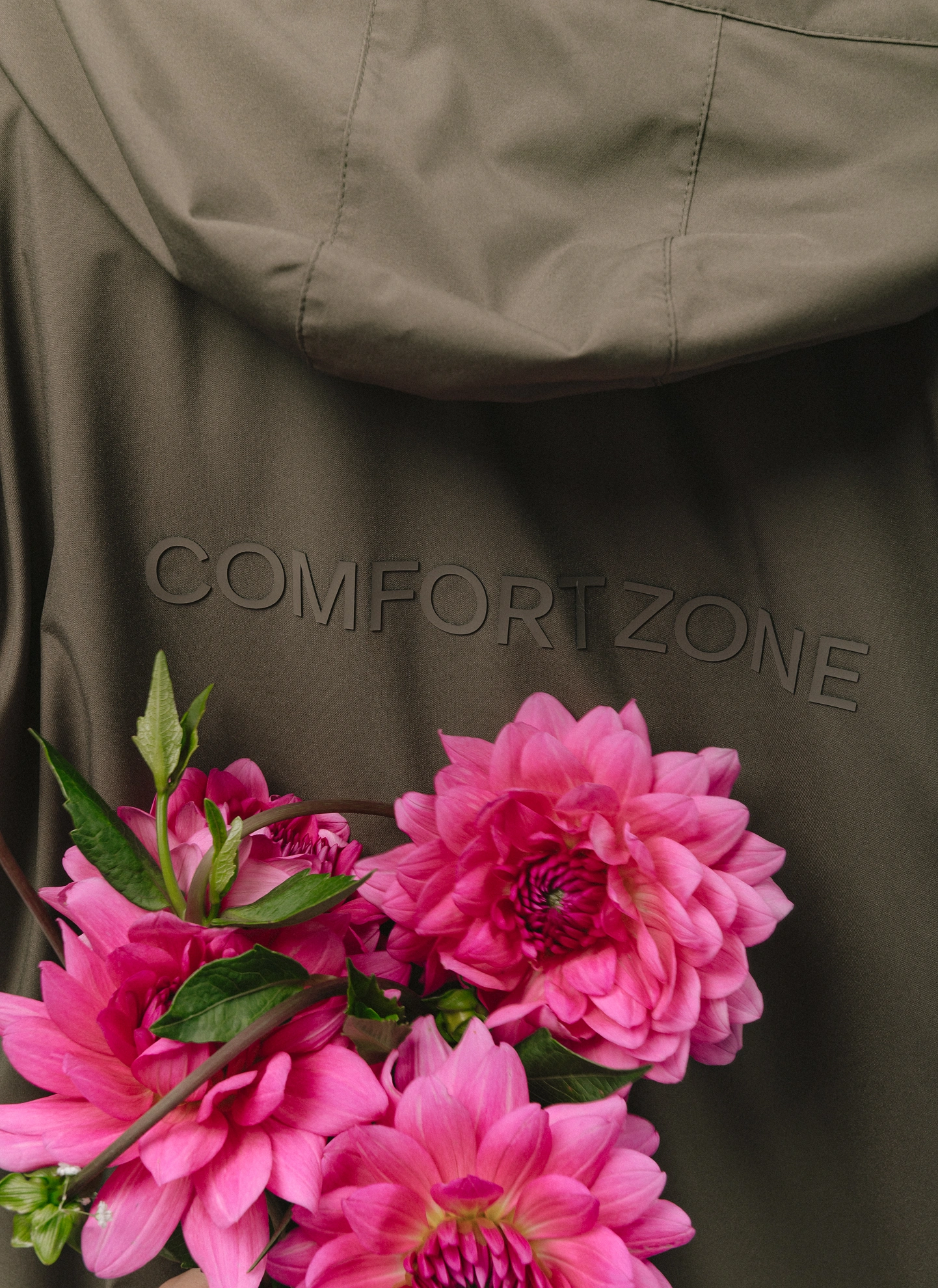 Zbliżenie na parkę przeciwdeszczową z kapturem w kolorze khaki z delikatnym napisem „Comfort Zone”, na tle której wyróżnia się bukiet wyrazistych, różowych dalii. Artystyczny detal z sesji zdjęciowej wzbogaca wizualnie lookbook Autumn Winter 2026 od Kuboty, akcentując połączenie estetyki z komfortem noszenia w chłodne dni.