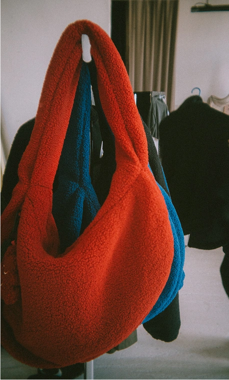 Trzy duże, pluszowe torby na ramię z miękkiego materiału typu fleece w kolorach czerwonym, kobaltowym i brązowym, zawieszone na stojaku odzieżowym. Ten wyrazisty detal akcesoriów wzbogaca oprawę wizualną lookbooka Autumn Winter 2026 Kuboty, prezentując przytulne i funkcjonalne dodatki do jesienno-zimowych stylizacji.